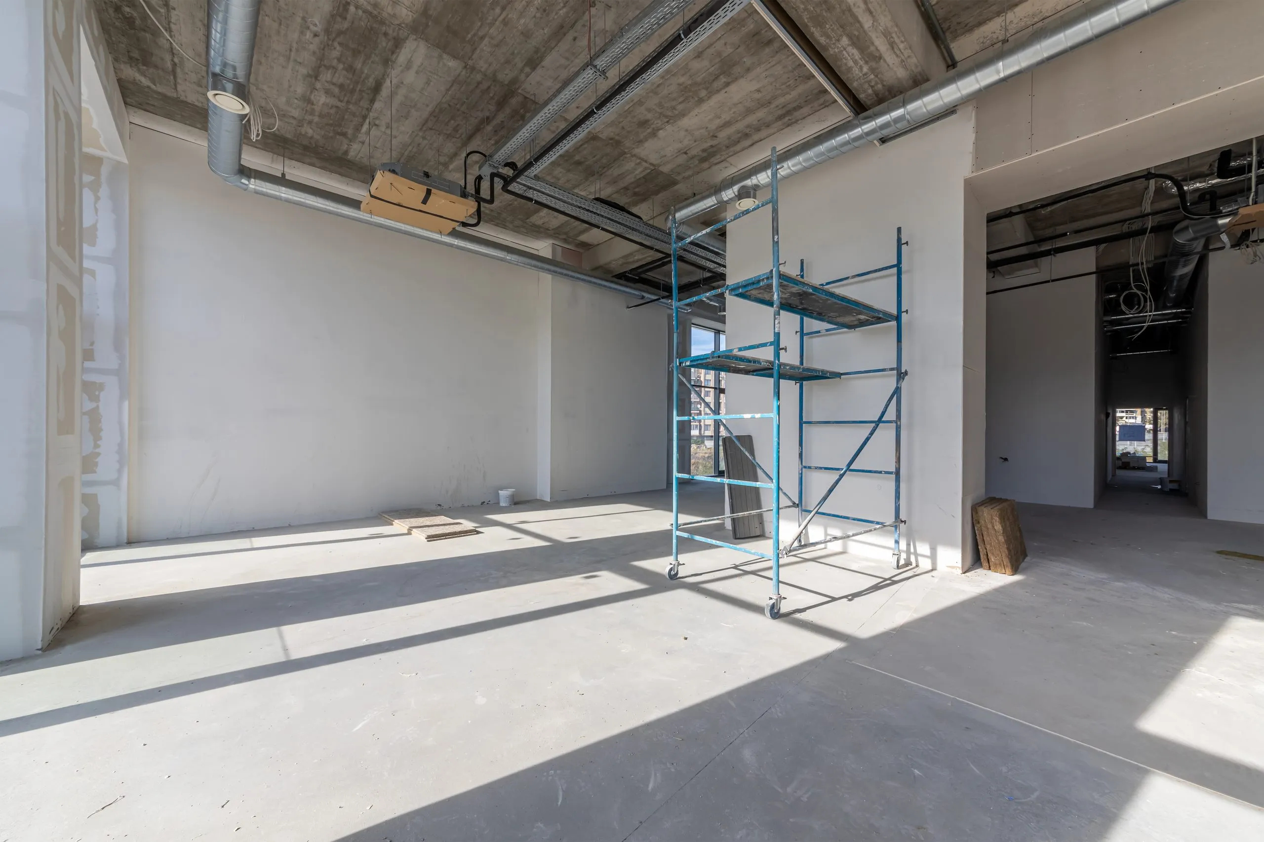 Empty commercial office suite mid-buildout, with taped and finished drywall partition walls, an exposed concrete ceiling carrying conduit and ductwork, and a blue mobile scaffold tower on a polished concrete floor