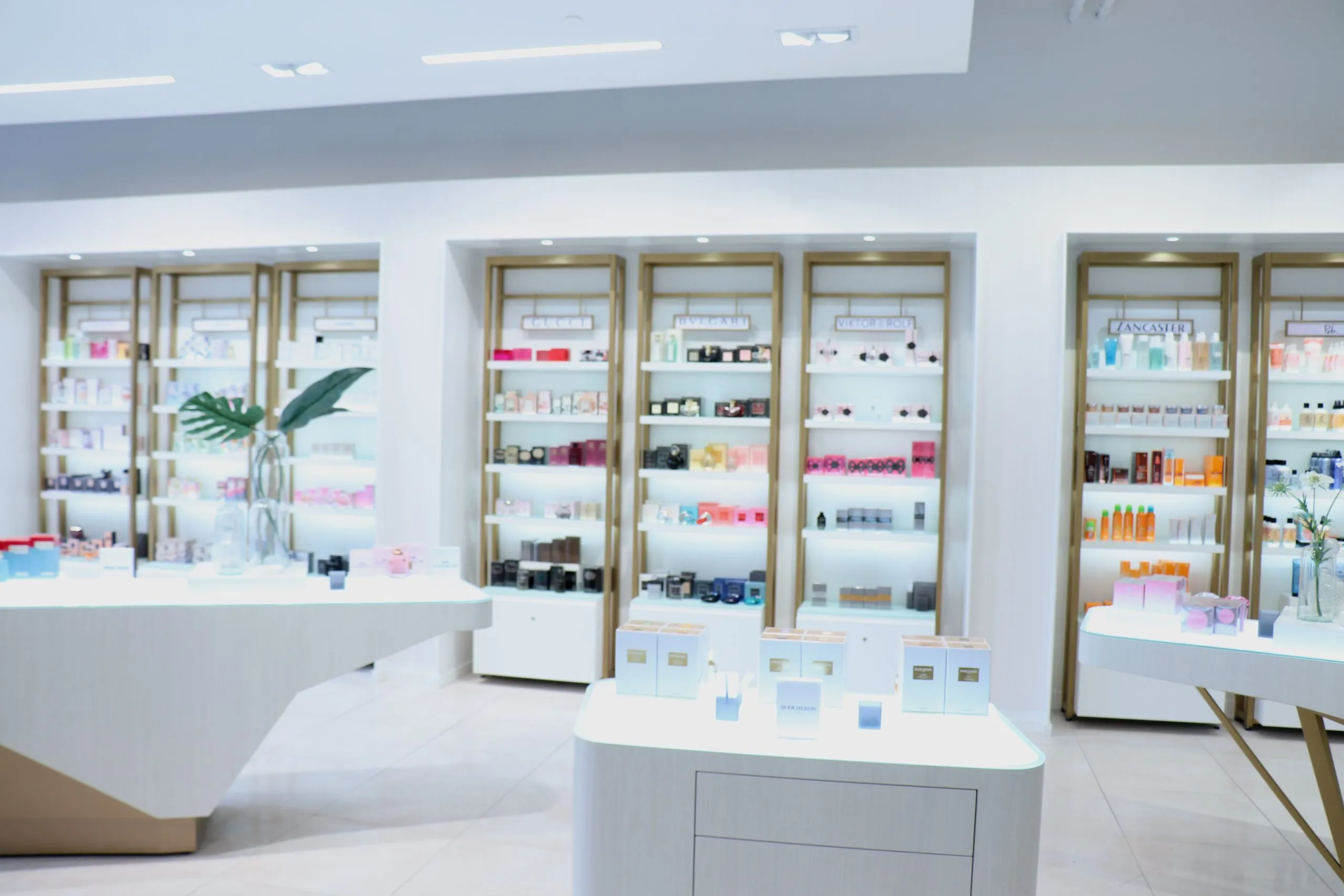 Finished upscale retail store interior with white display cabinetry, gold-trimmed internally lit product shelving, freestanding display tables, and a smooth painted ceiling with recessed downlights