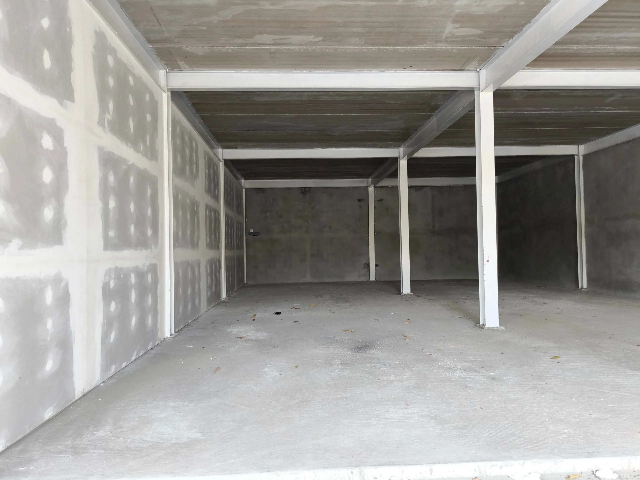 Empty industrial warehouse shell with white painted steel beams and columns, bare concrete walls, and a finished drywall partition wall showing taped and spotted joints, on a concrete floor